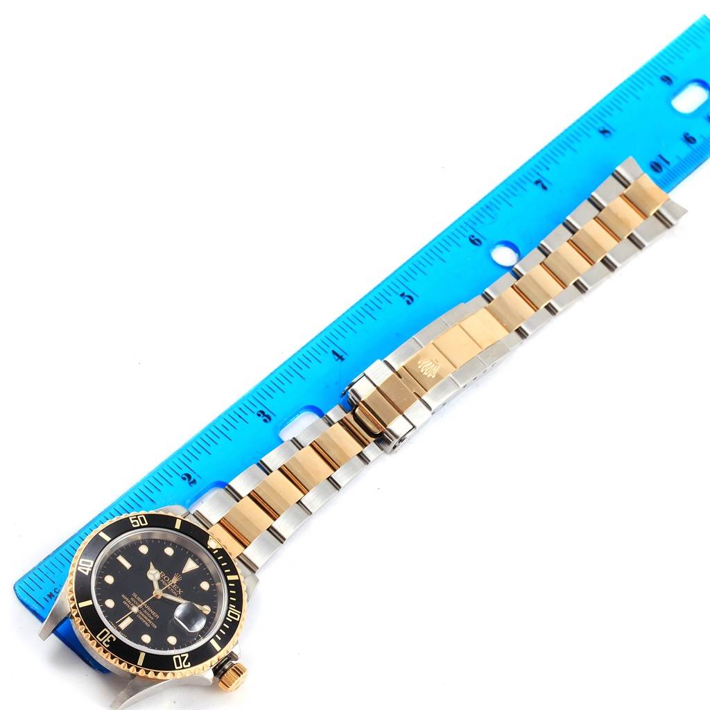 This image shows a Rolex Submariner 16613 Men
s Steel and Gold (two tone) Black Dial 16613 Men
s Steel and Gold (two tone) Black Dial watch from the top-down view, with its face, bezel, and metal bracelet alongside a blue ruler.