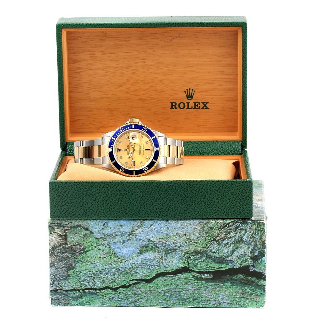 The image shows a Rolex Submariner 16613 Men
s Steel and Gold (two tone) Champagne Dial 16613 Men
s Steel and Gold (two tone) Champagne Dial watch with a gold dial and blue bezel, presented in an open green and wood Rolex box.