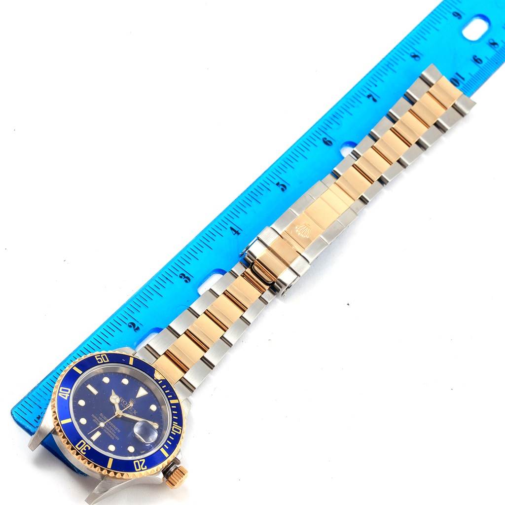 The image shows a Rolex Submariner 16613 Men's Steel and Gold (two tone) Blue Dial 16613 Men's Steel and Gold (two tone) Blue Dial watch laid flat next to a ruler, displaying its blue dial, bezel, and two-tone metallic bracelet.