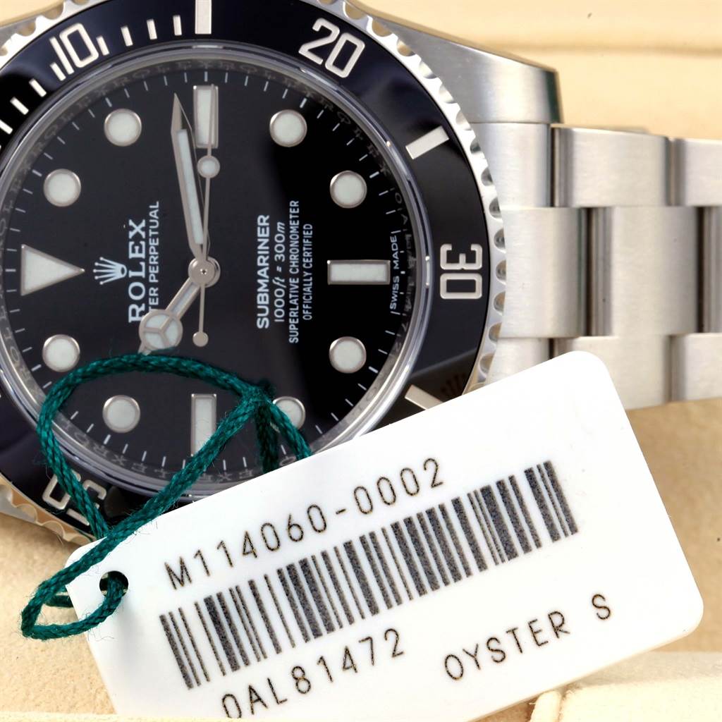 The image shows a close-up of the Rolex Submariner 114060 Men's Stainless Steel Black Dial 114060 Men's Stainless Steel Black Dial's face, bezel, and part of the bracelet, with a tag displaying model and serial numbers.