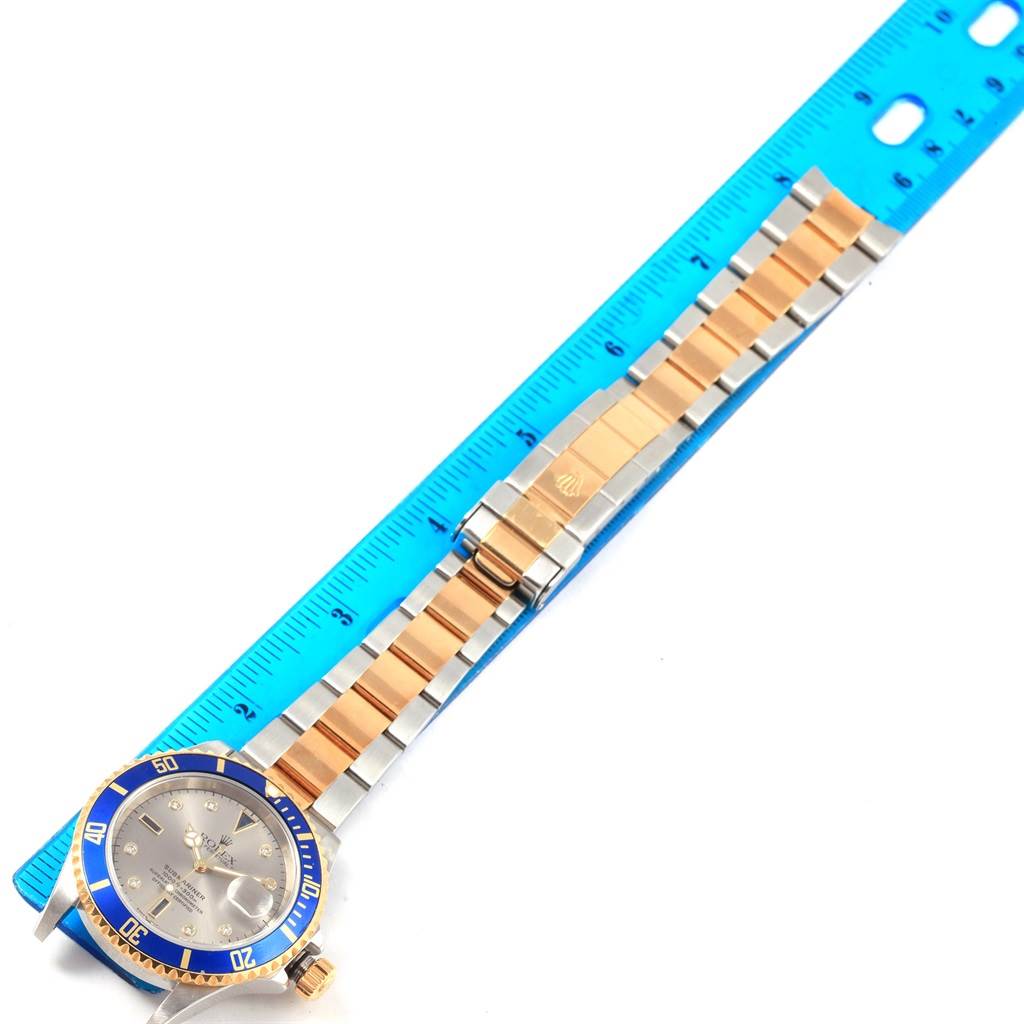 The image shows a Rolex Submariner 16613 Men
s Steel and Gold (two tone) Grey Dial 16613 Men
s Steel and Gold (two tone) Grey Dial watch laid flat with a blue ruler, highlighting the face, bezel, band, and clasp.