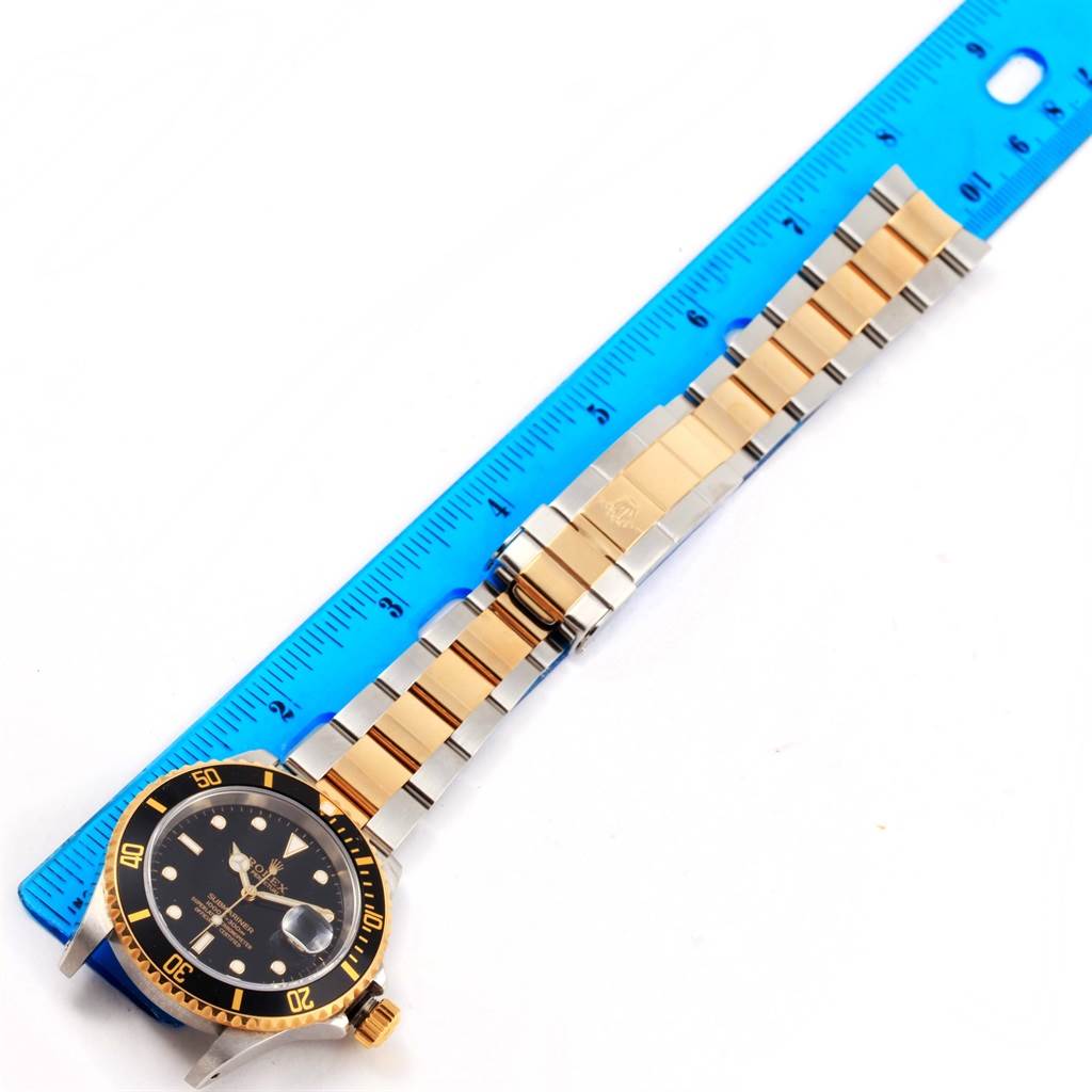 The Rolex Submariner 16613 Men's Steel and Gold (two tone) Black Dial 16613 Men's Steel and Gold (two tone) Black Dial watch is shown from a top-down angle, displaying the face, bezel, and bracelet alongside a blue ruler.