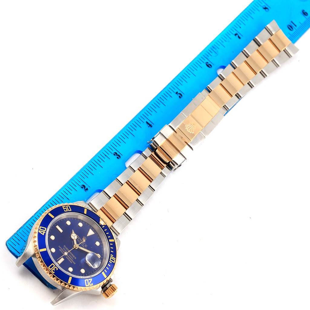 The Rolex Submariner 16613 Men's Steel and Gold (two tone) Blue Dial 16613 Men's Steel and Gold (two tone) Blue Dial watch is shown flat with its bracelet extended next to a blue ruler for scale.