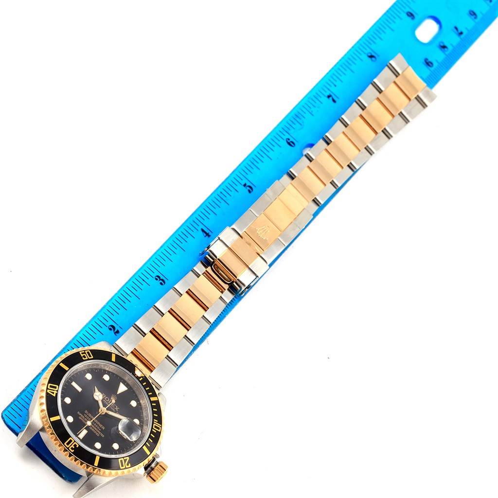The image shows a Rolex Submariner 16613 Men
s Steel and Gold (two tone) Black Dial 16613 Men
s Steel and Gold (two tone) Black Dial watch lying flat with its band extended alongside a blue ruler.