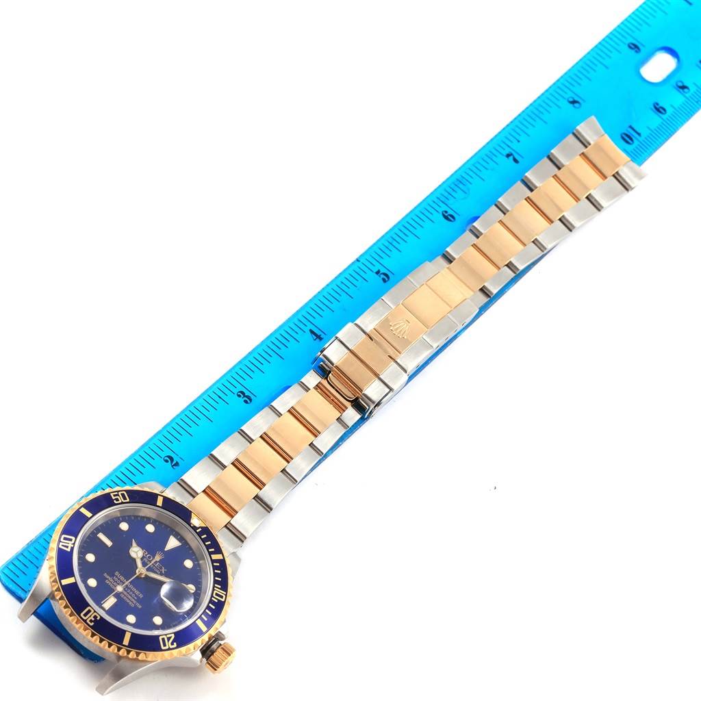 The image shows a Rolex Submariner 16613 Men's Steel and Gold (two tone) Blue Dial 16613 Men's Steel and Gold (two tone) Blue Dial watch with a blue dial and two-tone bracelet next to a ruler for measurement.