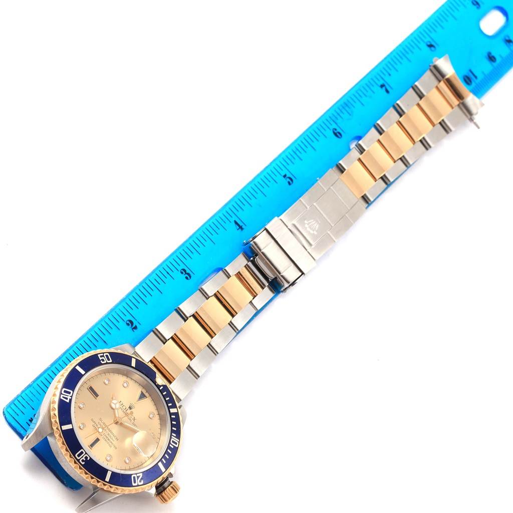 The image shows a Rolex Submariner 16613 Men's Steel and Gold (two tone) Champagne Dial 16613 Men's Steel and Gold (two tone) Champagne Dial watch laid flat with its bracelet and a blue ruler for scale.
