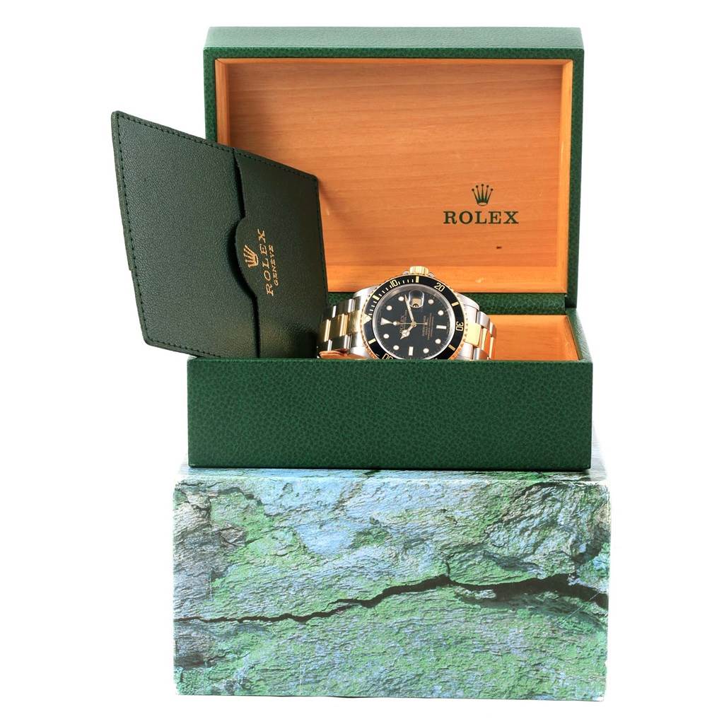 The image shows a Rolex Submariner 16613 Men
s Steel and Gold (two tone) Black Dial 16613 Men
s Steel and Gold (two tone) Black Dial watch in its open box, along with a green cardholder.