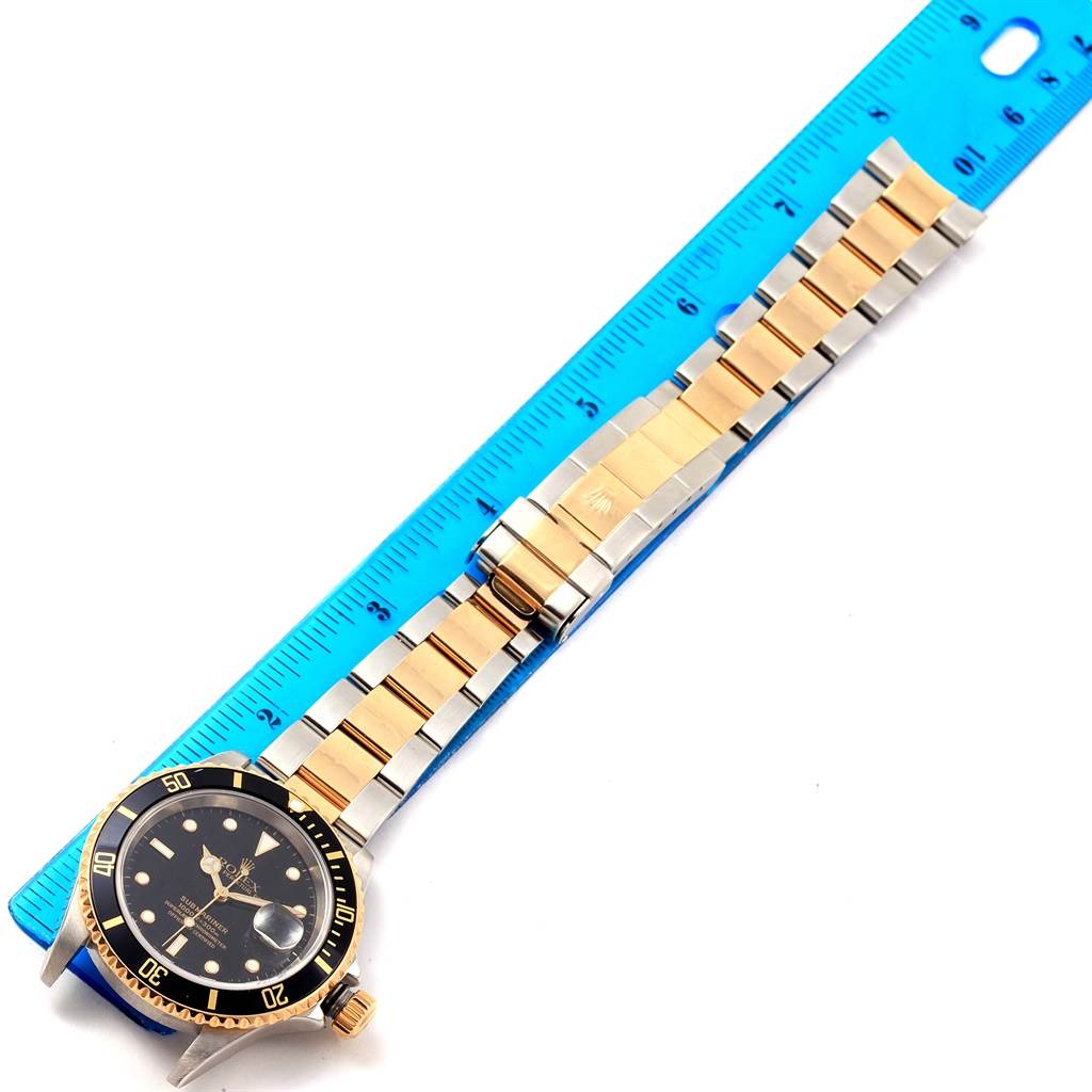 The image shows a full view of a Rolex Submariner 16613 Men
s Steel and Gold (two tone) Black Dial 16613 Men
s Steel and Gold (two tone) Black Dial watch placed next to a blue ruler for scale.