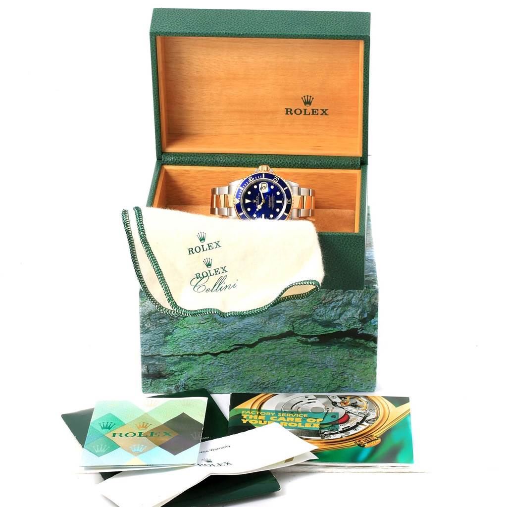 This image shows a Rolex Submariner 16613 Men's Steel and Gold (two tone) Blue Dial 16613 Men's Steel and Gold (two tone) Blue Dial watch inside an open Rolex box, along with branded booklet and a cleaning cloth.