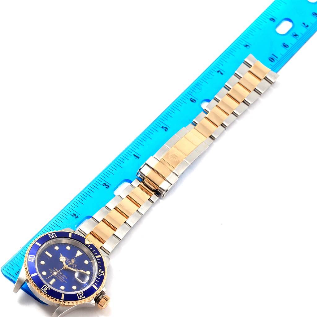 The Rolex Submariner 16613 Men's Steel and Gold (two tone) Blue Dial 16613 Men's Steel and Gold (two tone) Blue Dial watch is shown at an angled top view, highlighting its face and partially extended two-tone bracelet next to a ruler.