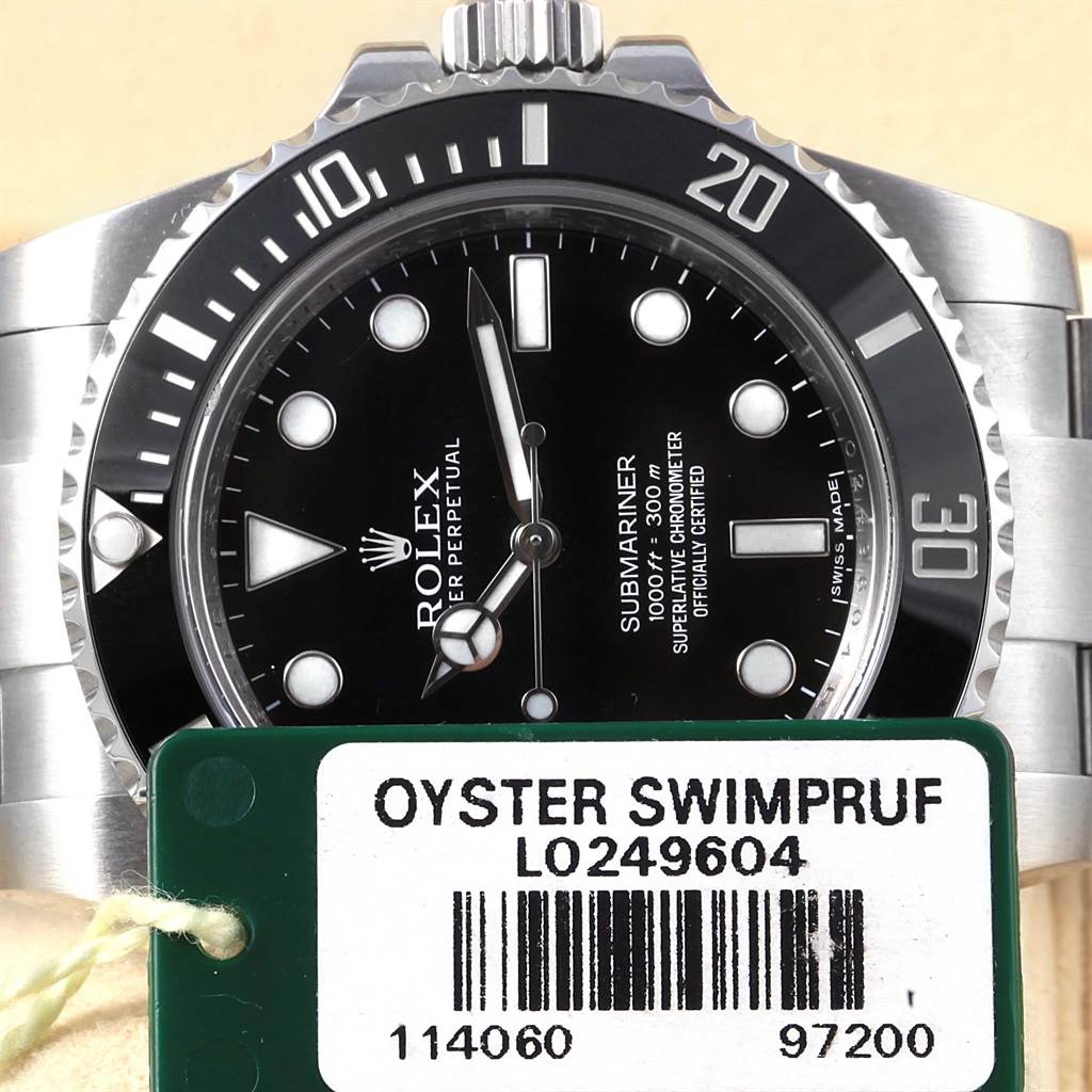 This image shows a close-up of the Rolex Submariner 114060 Men's Stainless Steel Black Dial 114060 Men's Stainless Steel Black Dial watch face and crown at an eye-level angle, with an Oyster Swimpruf tag.