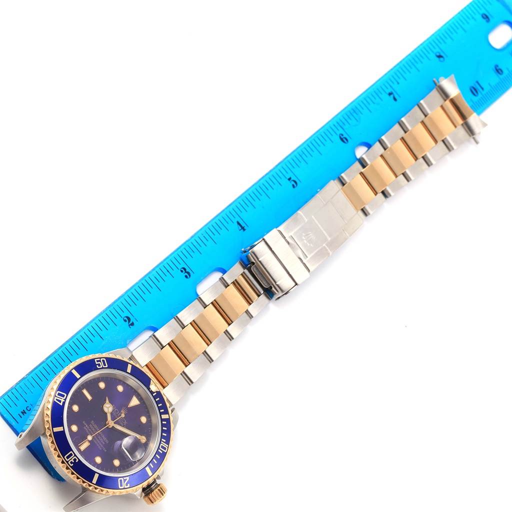 The Rolex Submariner 16613 Men
s Steel and Gold (two tone) Purple Dial 16613 Men
s Steel and Gold (two tone) Purple Dial watch is shown flat with a blue bezel and face, gold and silver bracelet, alongside a blue ruler for scale.