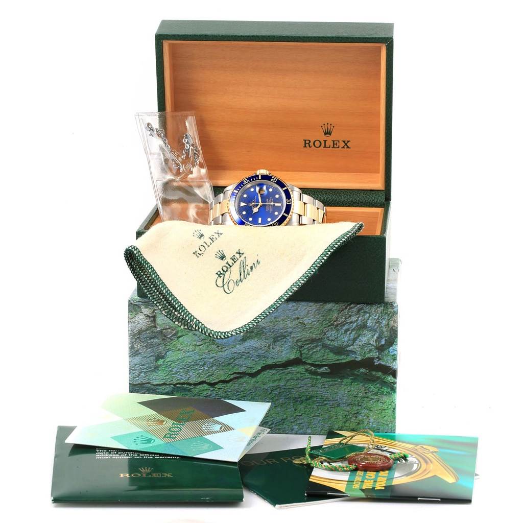 The image shows the Rolex Submariner 16613 Men's Steel and Gold (two tone) Blue Dial 16613 Men's Steel and Gold (two tone) Blue Dial watch displayed in an open case with accompanying booklets and a cleaning cloth.