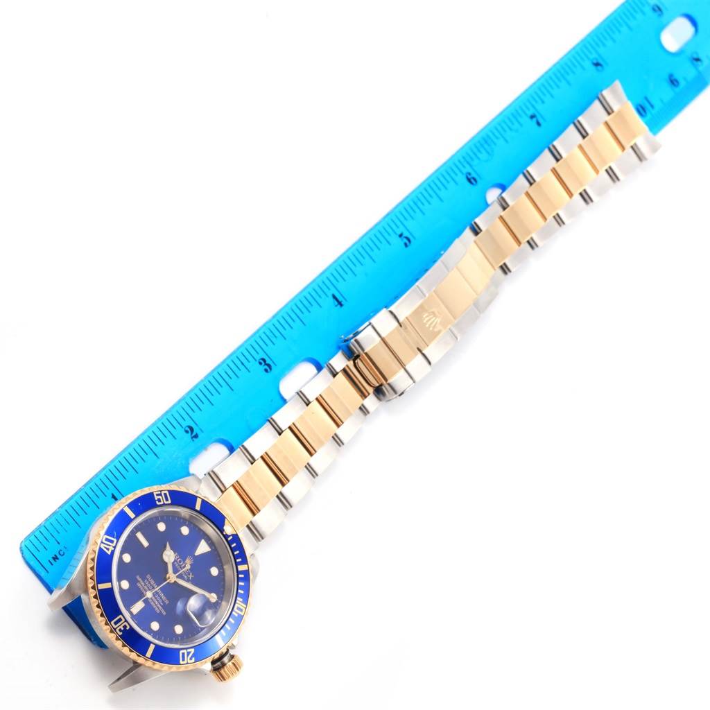 The image shows a Rolex Submariner 16613 Men's Steel and Gold (two tone) Blue Dial 16613 Men's Steel and Gold (two tone) Blue Dial watch laid flat next to a ruler, highlighting its dial, bezel, bracelet, and clasp.