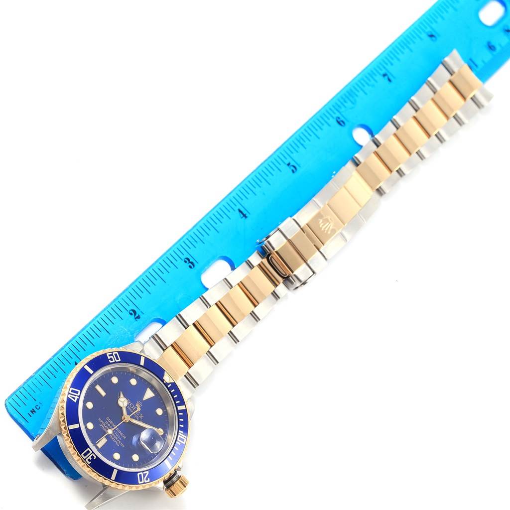 The image shows a Rolex Submariner 16613 Men's Steel and Gold (two tone) Blue Dial 16613 Men's Steel and Gold (two tone) Blue Dial watch laid flat next to a blue ruler, highlighting the face, bezel, and bracelet.
