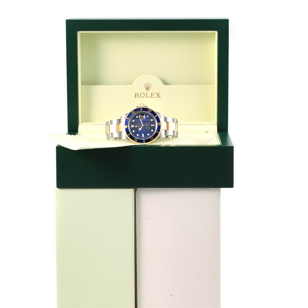 The Rolex Submariner 16613 Men's Steel and Gold (two tone) Blue Dial 16613 Men's Steel and Gold (two tone) Blue Dial watch is displayed front-facing in an open, green and cream-colored Rolex box.