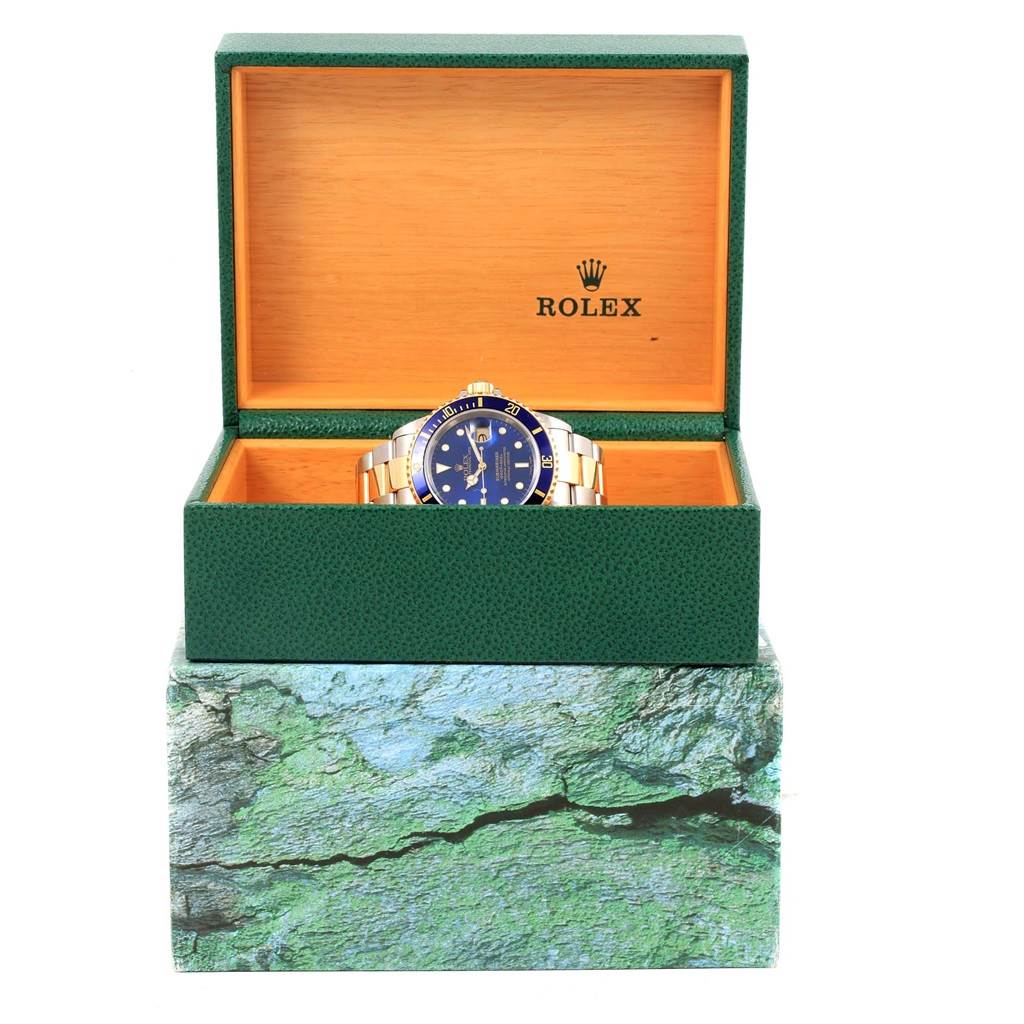 The image shows the front view of a Rolex Submariner 16613 Men's Steel and Gold (two tone) Blue Dial 16613 Men's Steel and Gold (two tone) Blue Dial watch in an open green and orange Rolex box.