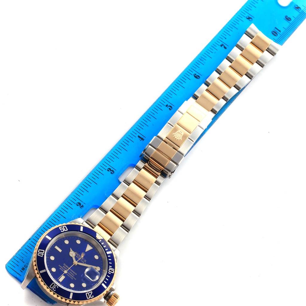 This is a Rolex Submariner 16613 Men's Steel and Gold (two tone) Blue Dial 16613 Men's Steel and Gold (two tone) Blue Dial watch laid flat with a blue ruler for scale, showing the full bracelet and face.