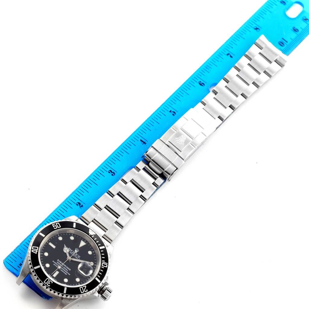 This image shows a full frontal view of a Rolex Submariner 16610 Men's Stainless Steel Black Dial 16610 Men's Stainless Steel Black Dial watch and its metal bracelet next to a blue ruler.