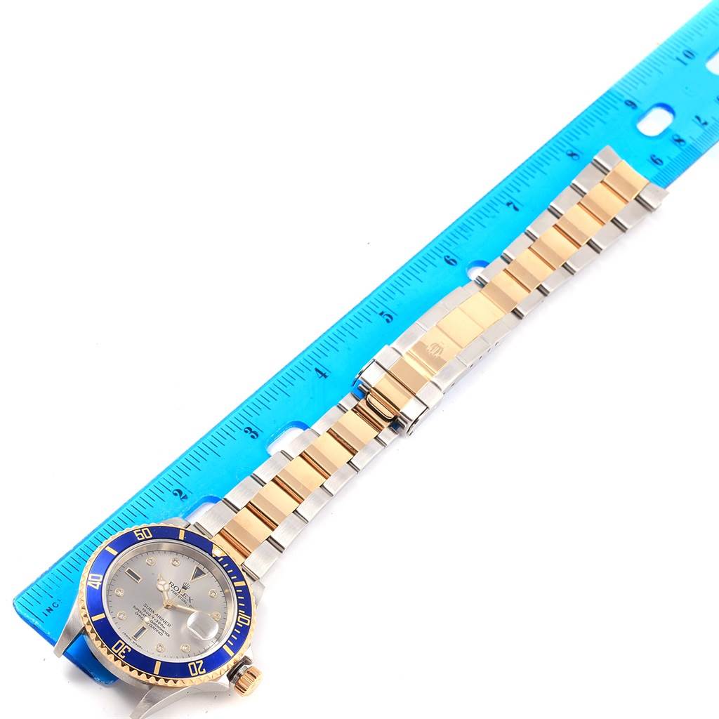 The image shows a Rolex Submariner 16613 Men's Steel and Gold (two tone) Grey Dial 16613 Men's Steel and Gold (two tone) Grey Dial watch with a blue bezel and a two-tone metal bracelet, displayed next to a blue ruler.