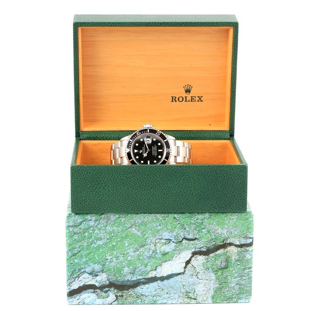 The Rolex Submariner 16610 Men's Stainless Steel Black Dial 16610 Men's Stainless Steel Black Dial watch is displayed front-on, partially inside its green and wooden display box.