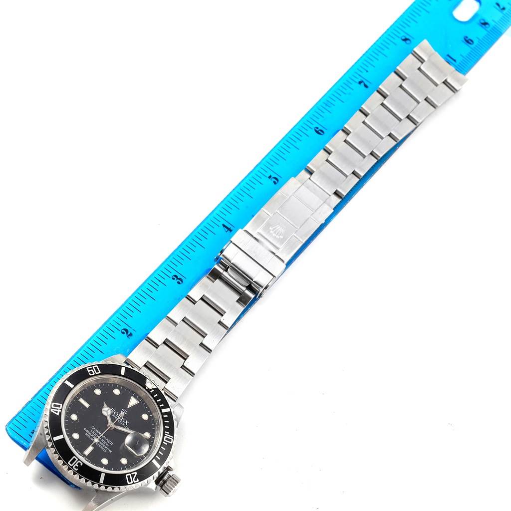 This image shows a Rolex Submariner 16610 Men's Stainless Steel Black Dial 16610 Men's Stainless Steel Black Dial watch placed alongside a blue ruler, highlighting its metal bracelet and face at a top-down angle.