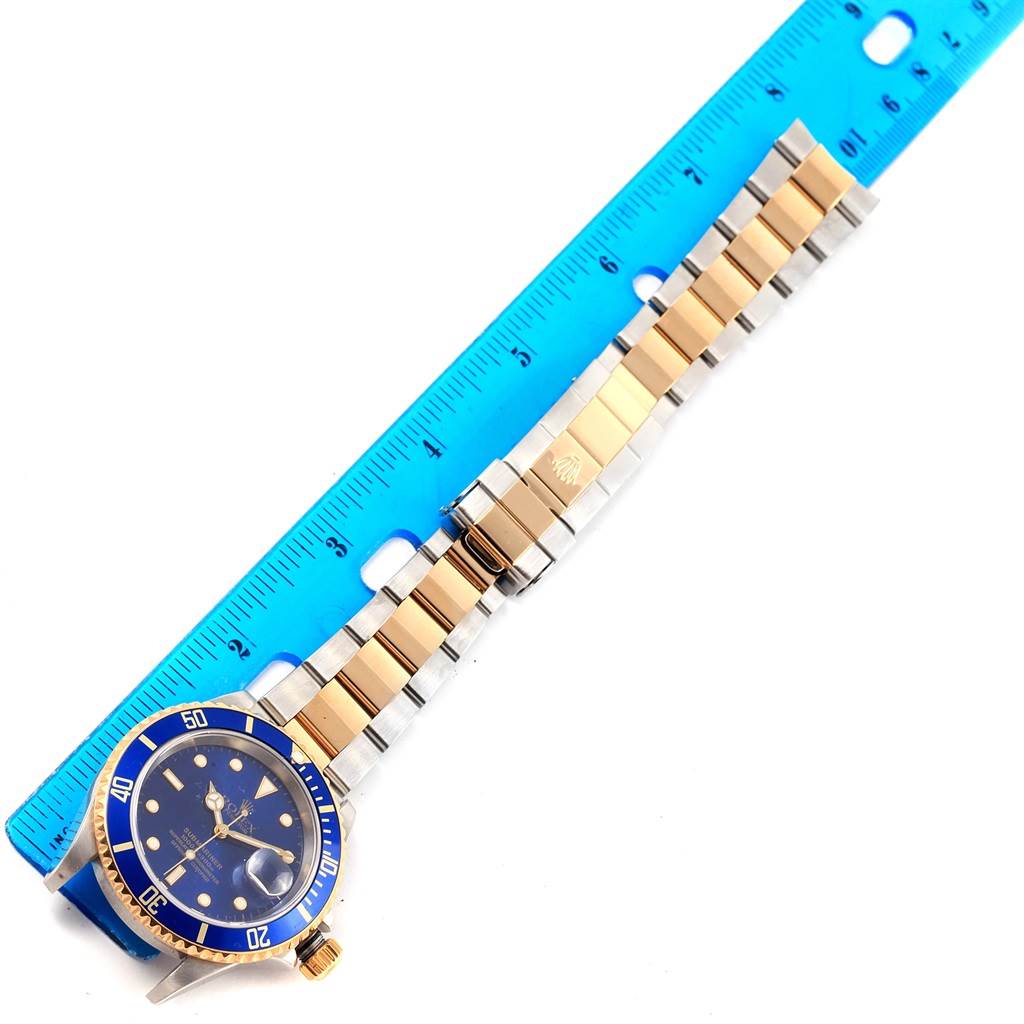 The image shows a top-down view of a Rolex Submariner 16613 Men's Steel and Gold (two tone) Blue Dial 16613 Men's Steel and Gold (two tone) Blue Dial, highlighting its bracelet, clasp, and blue bezel alongside a ruler.