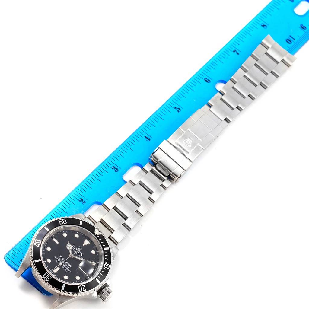 The Rolex Submariner 16610 Men's Stainless Steel Black Dial 16610 Men's Stainless Steel Black Dial watch is shown flat, displaying its face, bezel, bracelet, and clasp with a blue ruler for measurement.