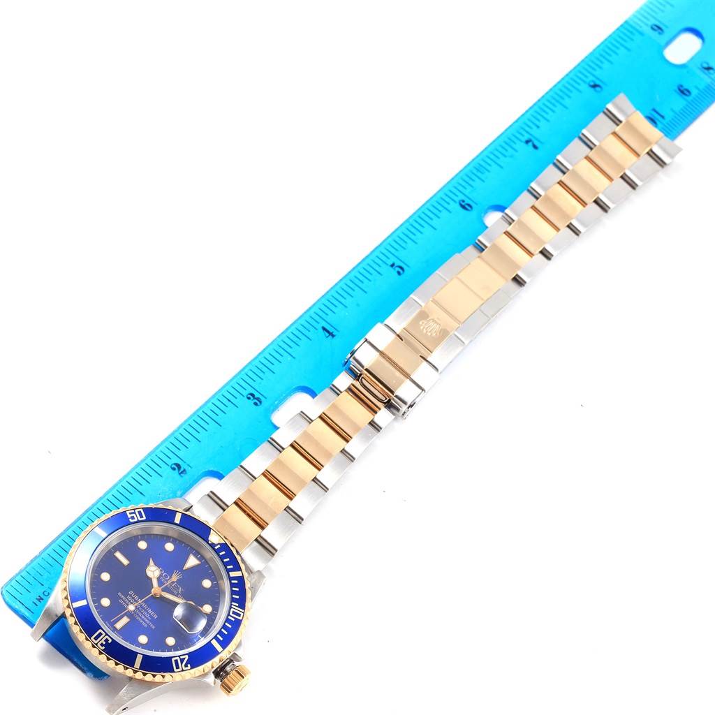 The image shows a Rolex Submariner 16613 Men's Steel and Gold (two tone) Blue Dial 16613 Men's Steel and Gold (two tone) Blue Dial watch laid flat next to a ruler, highlighting its gold and silver bracelet and blue dial.