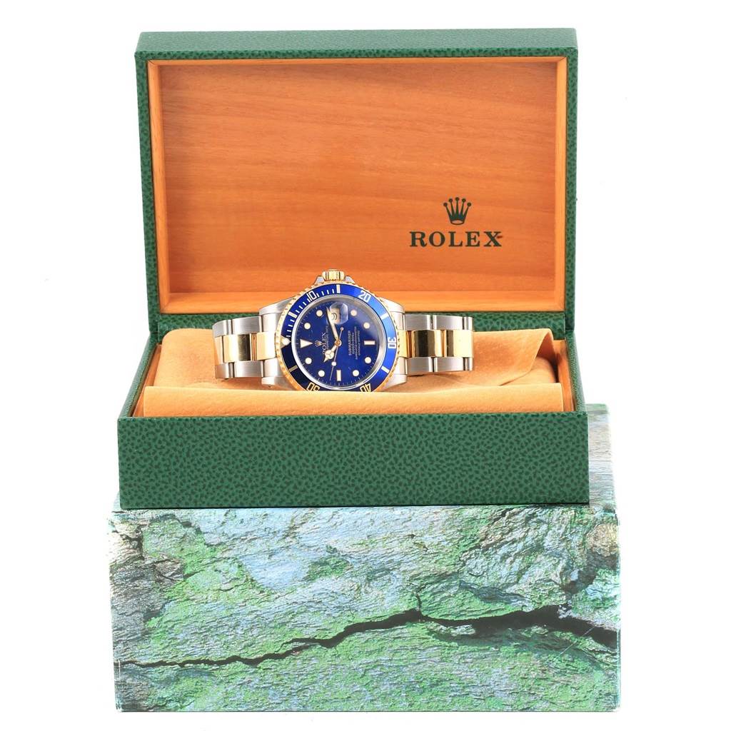 The Rolex Submariner 16613 Men's Steel and Gold (two tone) Blue Dial 16613 Men's Steel and Gold (two tone) Blue Dial watch is displayed front-facing in an open green and wooden box, showing the watch's face, bezel, and bracelet.