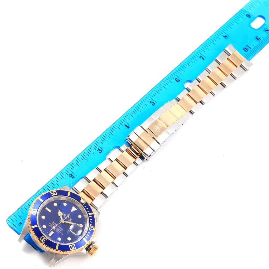 The image shows a Rolex Submariner 16613 Men's Steel and Gold (two tone) Blue Dial 16613 Men's Steel and Gold (two tone) Blue Dial watch with a blue dial and a two-tone bracelet, next to a blue ruler.