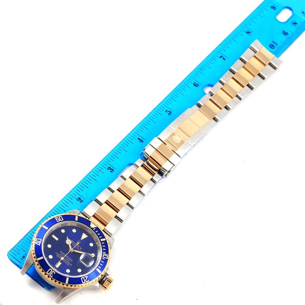 The image shows the front view of a Rolex Submariner 16613 Men's Steel and Gold (two tone) Blue Dial 16613 Men's Steel and Gold (two tone) Blue Dial with a blue dial and bezel, placed next to a ruler.