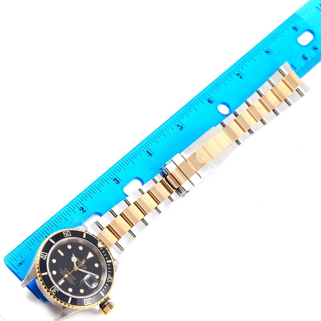 The image shows a Rolex Submariner 16613 Men's Steel and Gold (two tone) Black Dial 16613 Men's Steel and Gold (two tone) Black Dial watch laid flat, with a focus on its black dial, rotating bezel, and two-tone bracelet beside a blue ruler.