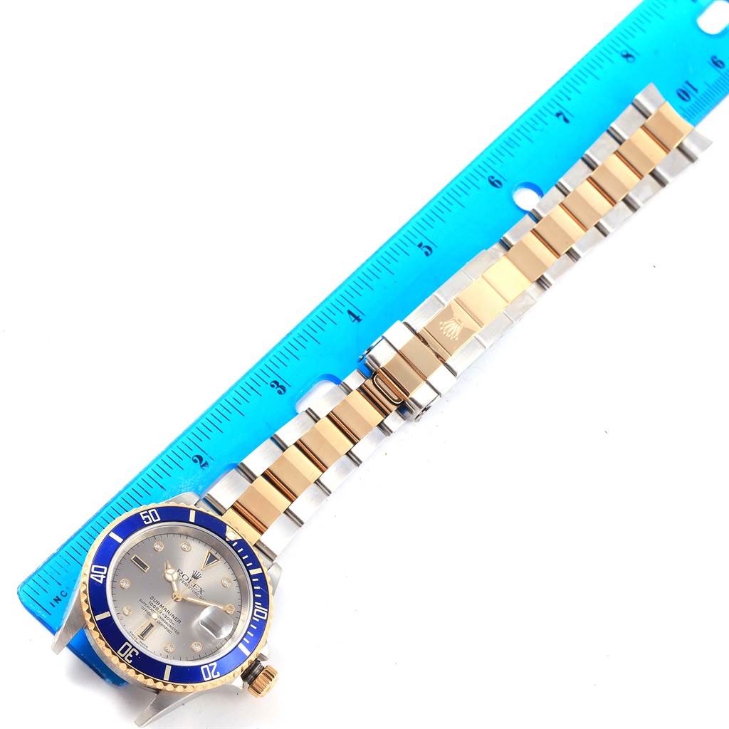 The image shows the face and bracelet of a Rolex Submariner 16613 Men's Steel and Gold (two tone) Grey Dial 16613 Men's Steel and Gold (two tone) Grey Dial watch with a blue bezel, next to a ruler.