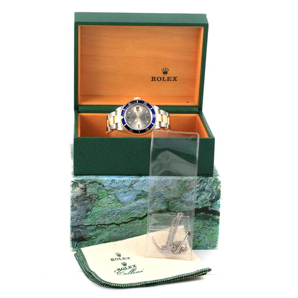 The Rolex Submariner 16613 Men's Steel and Gold (two tone) Grey Dial 16613 Men's Steel and Gold (two tone) Grey Dial watch is displayed front-on, inside its green box with an anchor accessory and a cleaning cloth placed outside.