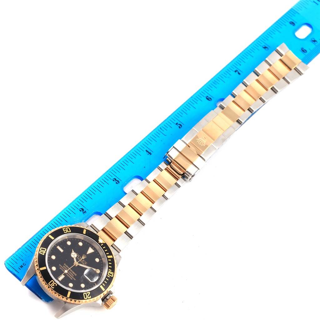 The image shows a Rolex Submariner 16613 Men's Steel and Gold (two tone) Black Dial 16613 Men's Steel and Gold (two tone) Black Dial watch laid flat next to a blue ruler, displaying the face and two-tone bracelet.