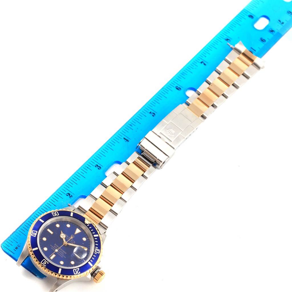 The image shows a Rolex Submariner 16613 Men's Steel and Gold (two tone) Blue Dial 16613 Men's Steel and Gold (two tone) Blue Dial watch laid next to a blue ruler, displaying the face, bezel, bracelet, and clasp.