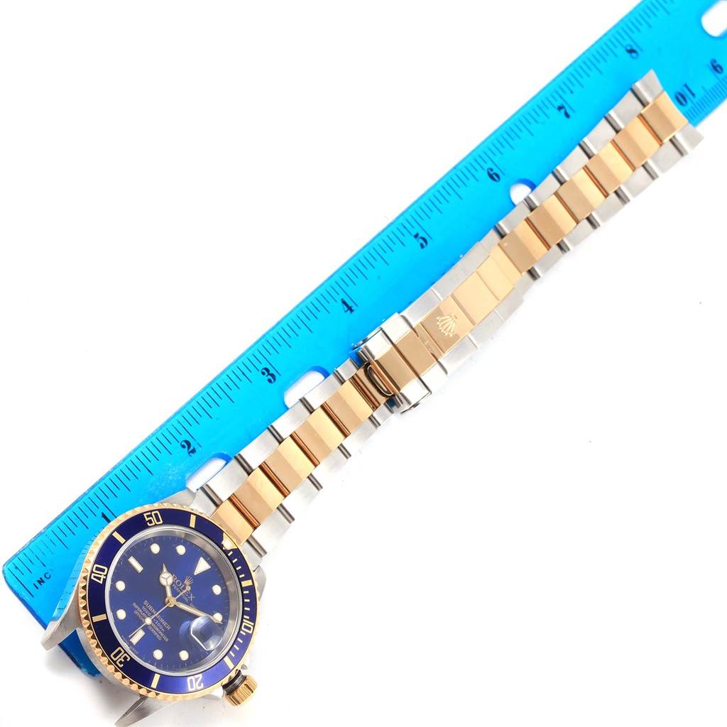 The Rolex Submariner 16613 Men's Steel and Gold (two tone) Blue Dial 16613 Men's Steel and Gold (two tone) Blue Dial watch is shown from a top-down angle, displaying its face, bezel, and bracelet alongside a blue ruler.