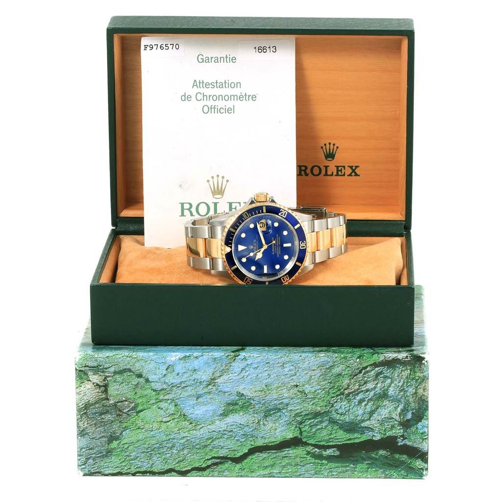 The image shows a Rolex Submariner 16613 Men's Steel and Gold (two tone) Blue Dial 16613 Men's Steel and Gold (two tone) Blue Dial watch with a blue dial and bezel, displayed upright in an open green box with a warranty card.