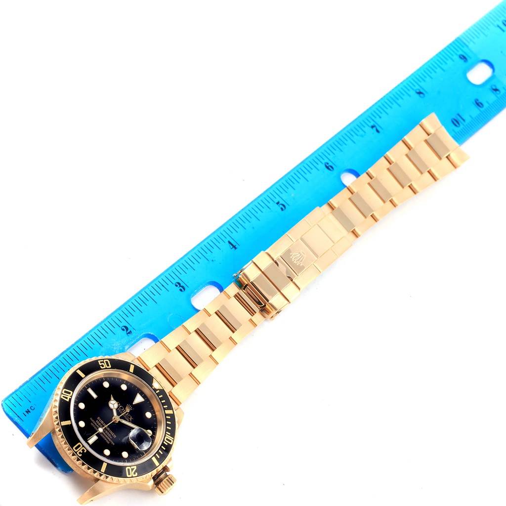 This image shows a Rolex Submariner 16618 Men
s Yellow Gold Black Dial 16618 Men
s Yellow Gold Black Dial watch laid flat with its bracelet and a blue ruler for scale.