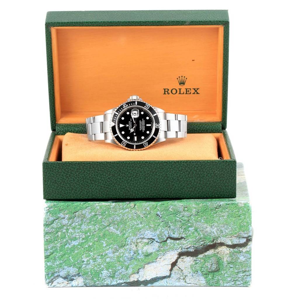 The Rolex Submariner 16610 Men's Stainless Steel Black Dial 16610 Men's Stainless Steel Black Dial watch is displayed front-facing in an open green box with an orange interior.