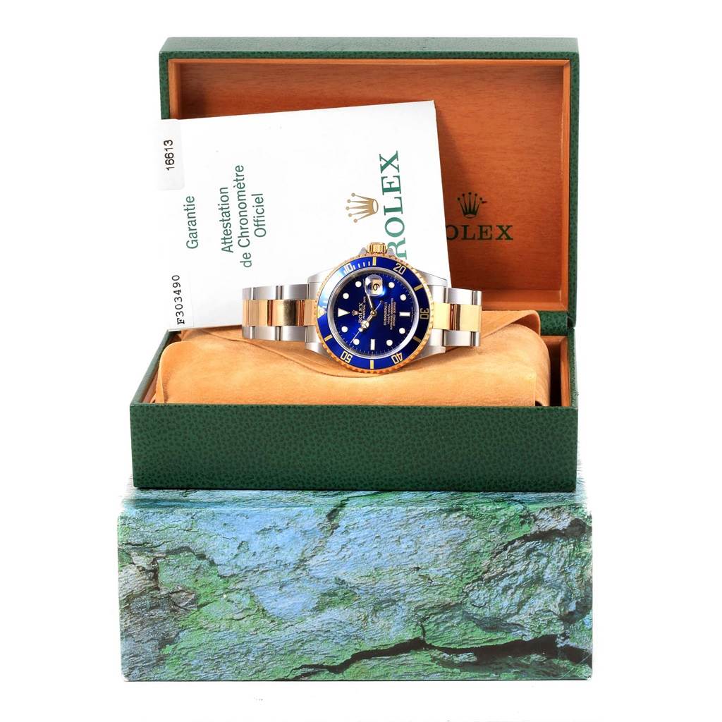 This is a front view of a Rolex Submariner 16613 Men's Steel and Gold (two tone) Blue Dial 16613 Men's Steel and Gold (two tone) Blue Dial watch, shown in its open display box with some documentation.