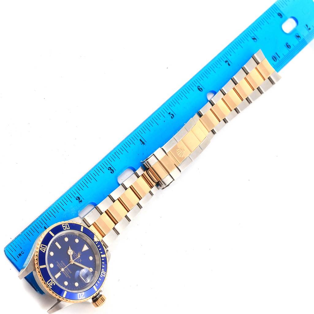 The image shows the front view of a Rolex Submariner 16613 Men's Steel and Gold (two tone) Blue Dial 16613 Men's Steel and Gold (two tone) Blue Dial watch and its bracelet measured against a blue ruler.