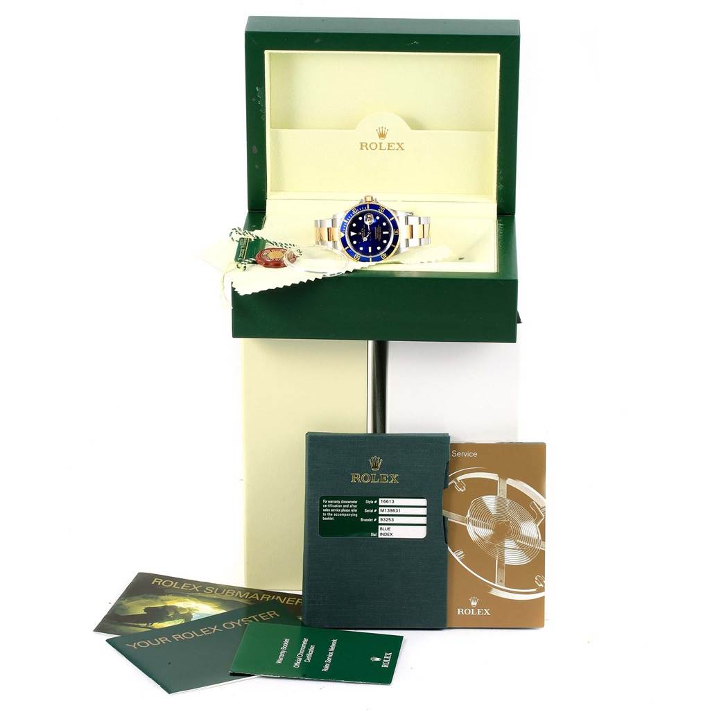 The image shows a Rolex Submariner 16613 Men's Steel and Gold (two tone) Blue Dial 16613 Men's Steel and Gold (two tone) Blue Dial watch at an angle, inside an open box with accompanying booklets and tags.