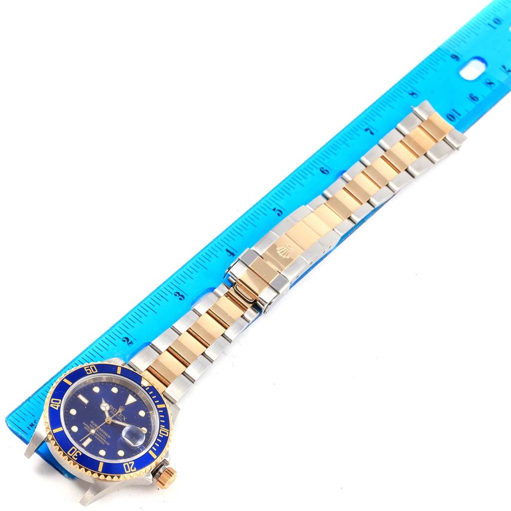 This image shows a Rolex Submariner 16613 Men's Steel and Gold (two tone) Blue Dial 16613 Men's Steel and Gold (two tone) Blue Dial watch laid flat next to a blue ruler, displaying the watch face and full bracelet.