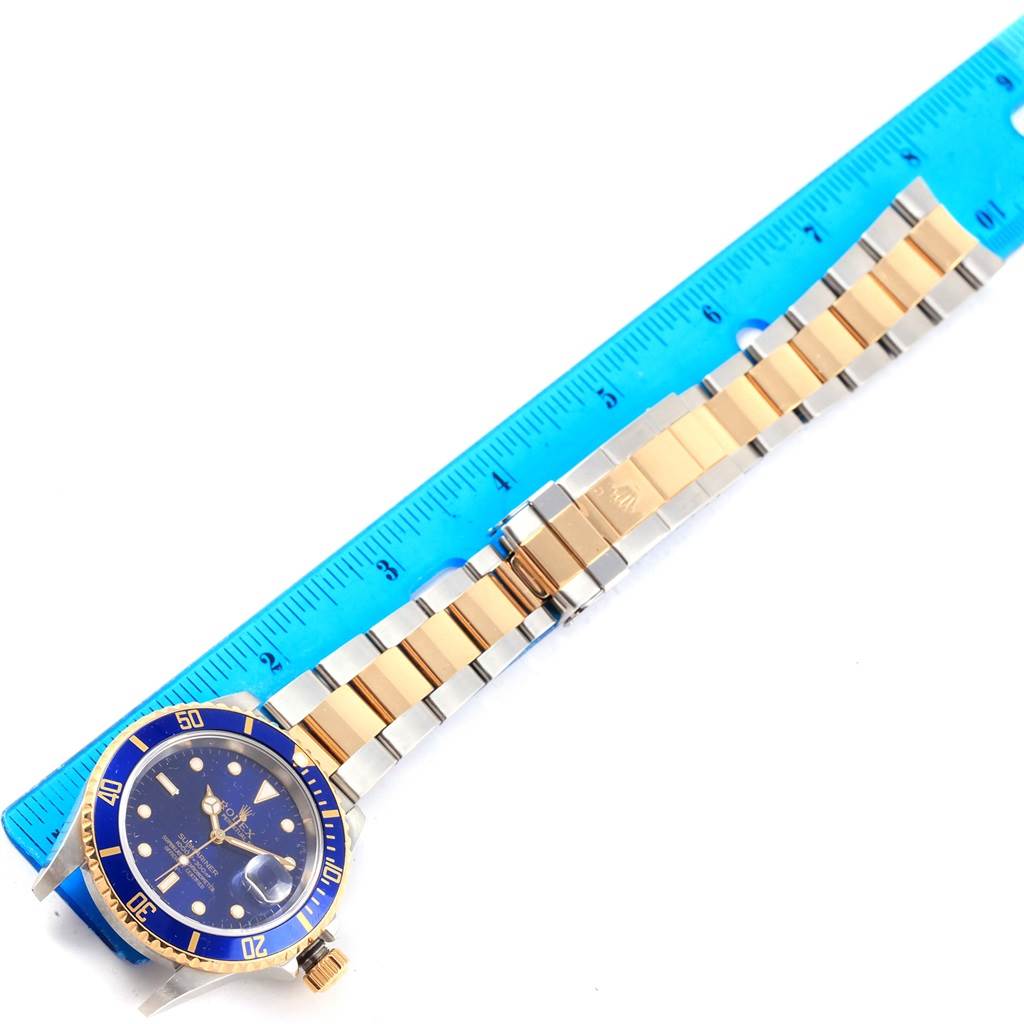 This image shows a Rolex Submariner 16613 Men's Steel and Gold (two tone) Blue Dial 16613 Men's Steel and Gold (two tone) Blue Dial watch laid flat next to a blue ruler, displaying its face, bezel, and bracelet.