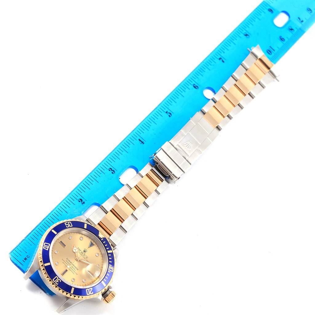 The Rolex Submariner 16613 Men's Steel and Gold (two tone) Champagne Dial 16613 Men's Steel and Gold (two tone) Champagne Dial watch is shown lying flat with the bracelet extended and aligned alongside a blue ruler.