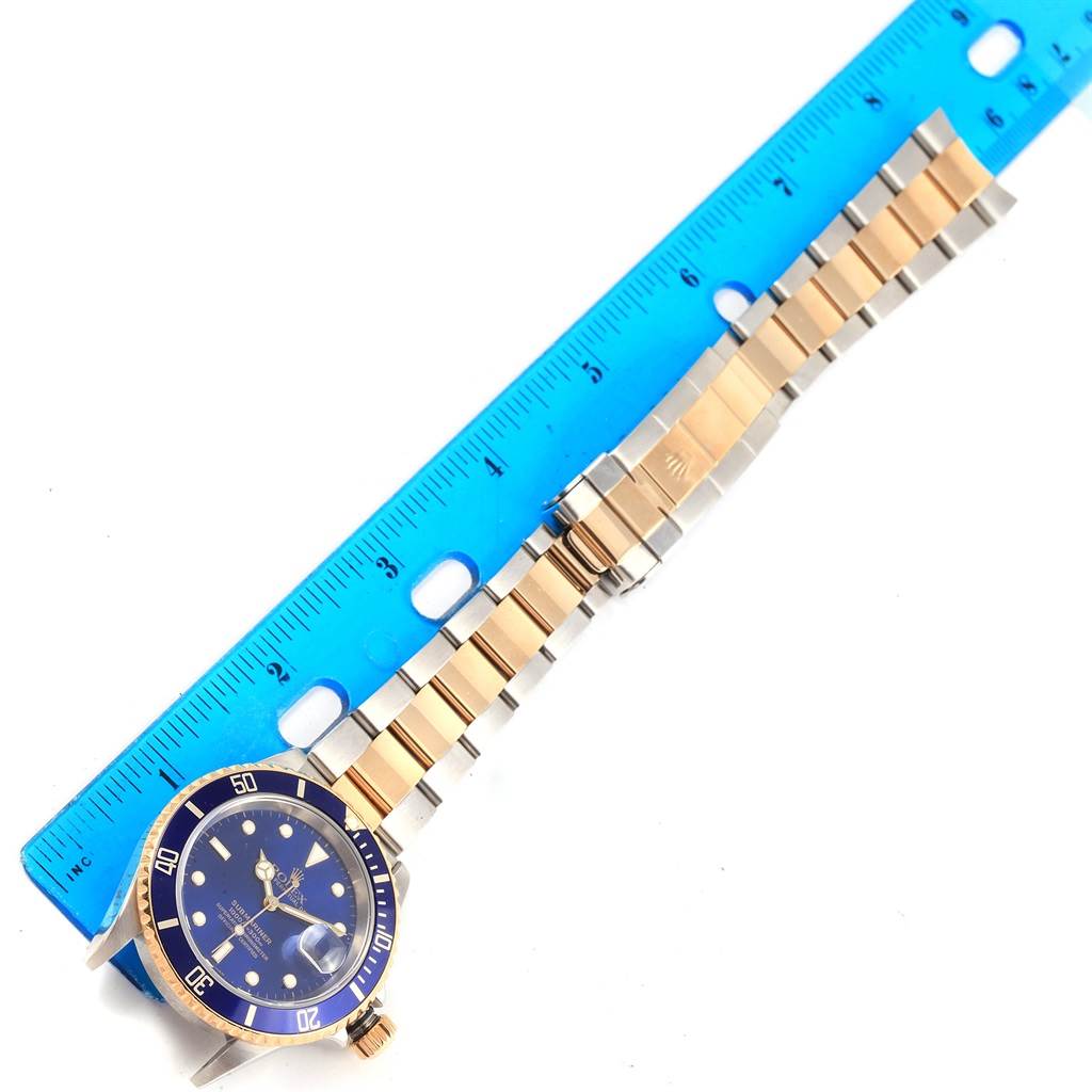 The image shows a top view of a Rolex Submariner 16613 Men's Steel and Gold (two tone) Blue Dial 16613 Men's Steel and Gold (two tone) Blue Dial watch with a blue dial and two-tone bracelet, placed next to a blue ruler.