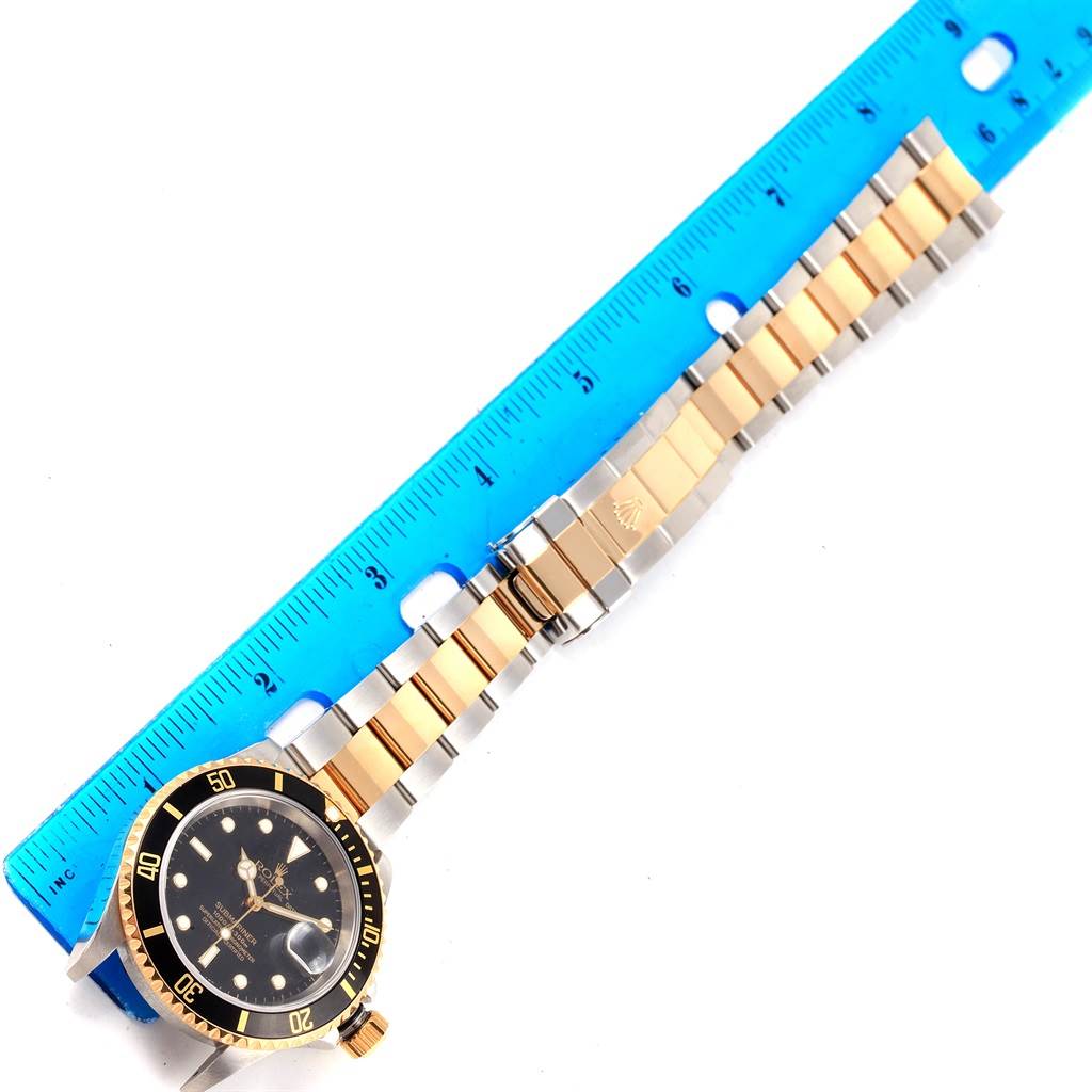 The image shows a Rolex Submariner 16613 Men's Steel and Gold (two tone) Black Dial 16613 Men's Steel and Gold (two tone) Black Dial watch laid flat with a blue ruler next to it, highlighting the face, bezel, and bracelet.
