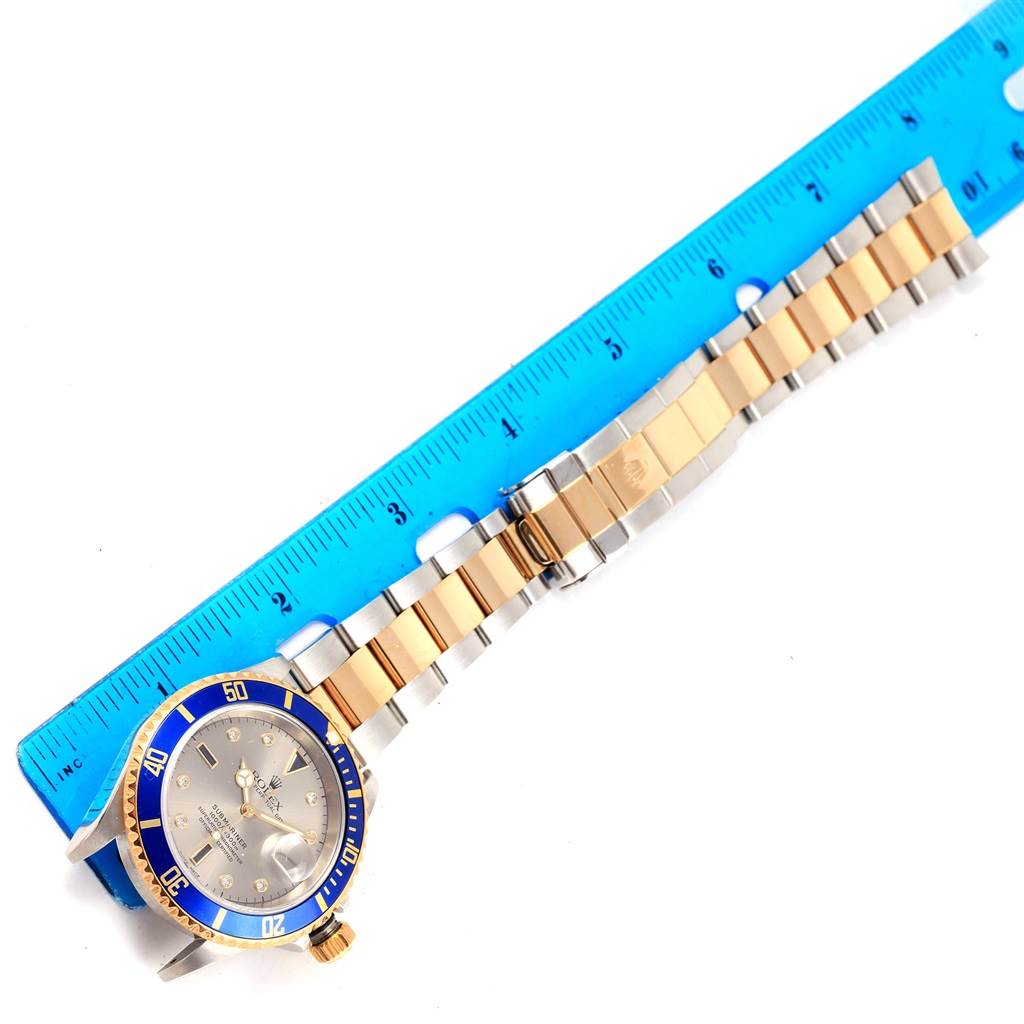 The Rolex Submariner 16613 Men's Steel and Gold (two tone) Grey Dial 16613 Men's Steel and Gold (two tone) Grey Dial watch is shown from a top-down angle, displaying its face, bracelet, and clasp alongside a blue ruler for scale.